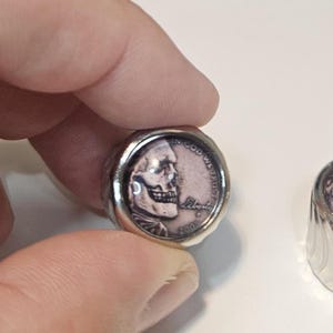 May include: A silver-toned ring featuring a circular inset made from a 2005 United States penny. The coin has been modified with a skull graphic overlaying the portrait, with the text IN GOD WE TRUST, LIBERTY, and the year 2005 clearly visible.
