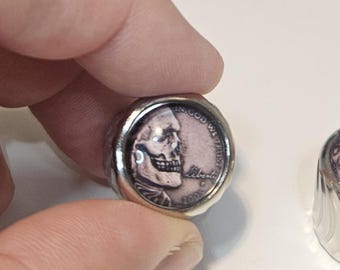 May include: A silver-toned ring featuring a circular inset made from a 2005 United States penny. The coin has been modified with a skull graphic overlaying the portrait, with the text IN GOD WE TRUST, LIBERTY, and the year 2005 clearly visible.
