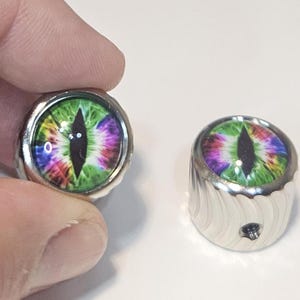 May include: Two cylindrical metal knobs featuring a vibrant, multi-colored eye graphic with a vertical black pupil. The top surface is domed and glossy, while the sides of the knobs have a textured, grooved pattern and a small threaded hole.
