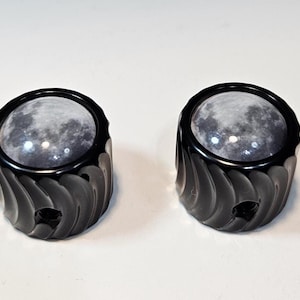 May include: Two black, decorative knobs with a spiral design. Each knob features a clear, circular insert displaying a detailed image of the moon. The knobs are set against a plain white background.