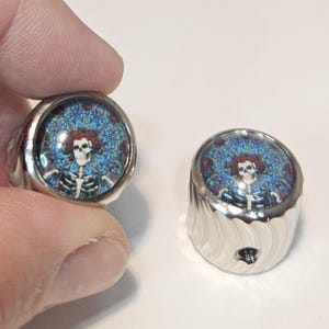May include: Two cylindrical silver metal knobs featuring a circular inlay on top. The inlay displays a colorful graphic of a skeleton with red hair against a blue and green mandala-style background. The sides of the knobs have a textured, grooved finish.