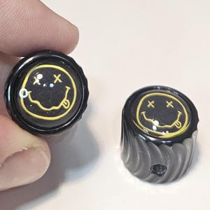 May include: Two black cylindrical control knobs featuring a yellow graphic of a smiley face with X-shaped eyes and a tongue sticking out. The knobs have a ribbed side texture and a circular top surface with a glossy finish and a set screw hole.