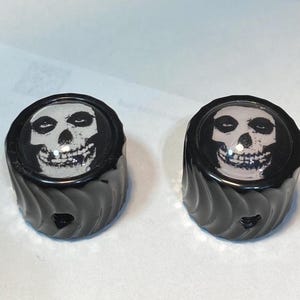 May include: Two black, cylindrical knobs with a skull graphic. The skull is white with black details, set against a black background. The knobs have a textured, swirled design on the sides.