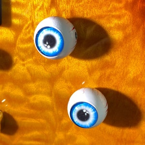 May include: Two white plastic eyeballs with blue irises and black pupils. The eyeballs are on a yellow and orange background.