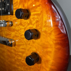 May include: Close-up of an electric guitar body with a flame maple finish in shades of amber and brown. Three black control knobs are visible, along with a portion of the bridge and pickups.
