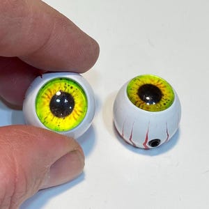 May include: Two realistic, white-based eyeballs with detailed yellow and black irises. One is held by a hand, the other rests on a white surface. The eyeballs have red veins and a black pupil, perfect for Halloween or craft projects.