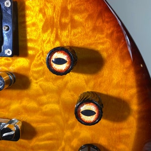 May include: Close-up of a guitar with two knobs. The knobs are black with a red and orange eye design.
