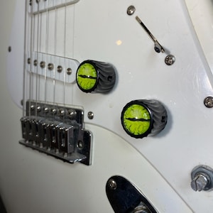 May include: A white electric guitar body featuring two custom control knobs with a black fluted edge and a bright green reptilian eye graphic with a vertical black pupil centered on the top of each knob, positioned near the bridge and pickups.