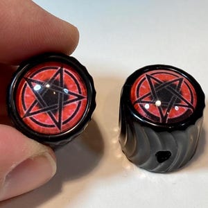 May include: Two black knobs with a red and black pentagram design. The knobs have a glossy finish and a ridged edge. One knob is held by a hand, and the other is resting on a surface.