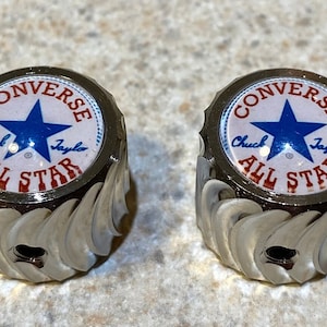 May include: Two silver-colored metal knobs with a white background and red and blue Converse All Star logo. The logo features a blue star and the text "Converse Chuck Taylor All Star".