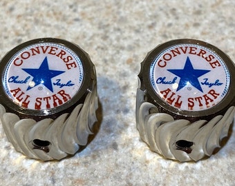 May include: Two silver-colored metal knobs with a white background and red and blue Converse All Star logo. The logo features a blue star and the text "Converse Chuck Taylor All Star".