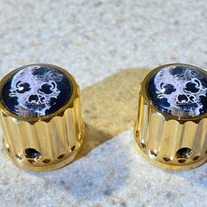 May include: Two gold-colored knobs with a skull design. The knobs have a ridged, cylindrical shape and a black and white skull image on top. The skull image is set against a black background.