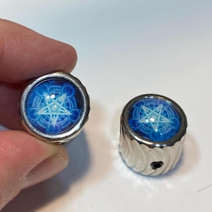 May include: Two silver-toned, cylindrical objects with a blue, circular design featuring a white pentagram. The design is set against a background of concentric circles and celestial patterns. The objects appear to be decorative or functional.