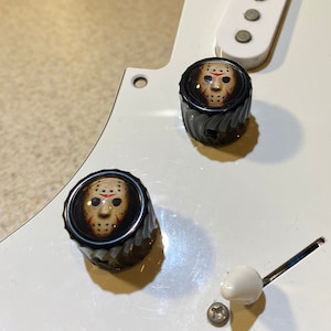 May include: Two black guitar knobs with a white background. The knobs have a graphic of a hockey mask with a red slash across the eyes.