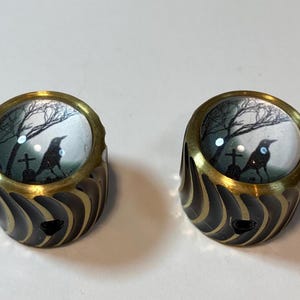May include: Two matching rings with a brass-colored rim and a black and gold patterned band. Each ring features a miniature scene of a crow perched on a tombstone under a bare tree, set against a cloudy sky.