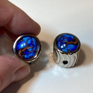 May include: Two silver rings with a vibrant blue and brown stone inlay. One ring is held by a hand, showcasing the intricate details of the stone's pattern. The other ring sits on a white surface.