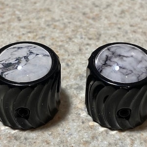 May include: Two black guitar knobs with a white and gray marbled design on the top. The knobs have a textured, spiral design.