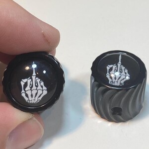 May include: Two black knobs with a white skeleton hand giving the middle finger. One knob is held by a hand. The other knob is on a white surface. The knobs have a ridged design.