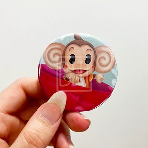 May include: A round button featuring a cartoon monkey with large ears, wearing a red and yellow outfit, and riding a red and pink boat. The monkey has a surprised expression and is holding onto the boat with its hands.