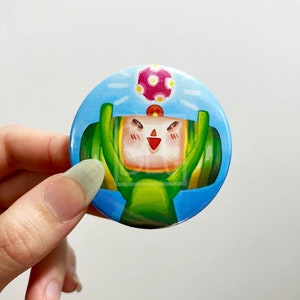 May include: A round button with a blue background and a cartoon character wearing a green helmet and holding a pink and white ball. The character has a smiling face with red cheeks.