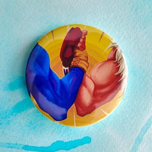 May include: A round button with a yellow background featuring two muscular arms in a handshake. One arm is blue, the other is red, and they are clasped together. The button has a glossy finish and is set against a light blue background.