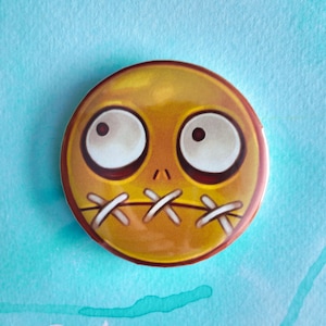 May include: A round, yellow button with a cartoon face. The face has large, white eyes with dark pupils, a small nose, and a stitched-up mouth. The button has a red border.