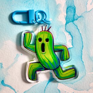 May include: A green and yellow cactus-shaped keychain with a surprised expression, featuring a blue clasp. The keychain is made of clear acrylic and has a cartoon-like design. The background is a watercolor wash of blue and white.