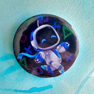 May include: Round button with a black background featuring a white and blue robot character with a smiling face. The robot has blue accents and is surrounded by colorful PlayStation controller symbols. The button is approximately 1.5 inches in diameter.