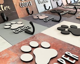 Op de afbeelding: Een set houten hondenriemhaken met pootprintontwerpen en gepersonaliseerde namen. De haken zijn in verschillende kleuren geverfd, waaronder bruin, grijs en wit. De namen op de haken zijn "Dexter", "Cooper", "Chico" en "Remi".
