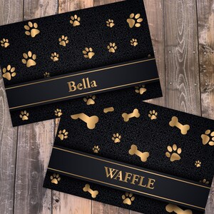 May include: Two rectangular cards featuring a black background with a dark, intricate filigree pattern. The cards are decorated with gold paw prints and bone shapes. Each card has a horizontal black band with the gold text Bella or WAFFLE.