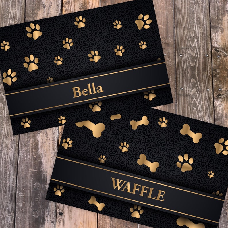 May include: Two rectangular cards featuring a black background with a dark, intricate filigree pattern. The cards are decorated with gold paw prints and bone shapes. Each card has a horizontal black band with the gold text Bella or WAFFLE.