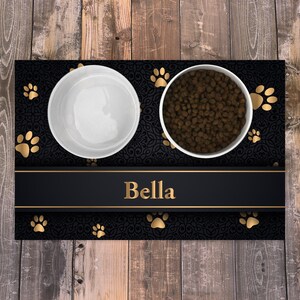 May include: A rectangular pet feeding mat featuring a black background with a dark ornate pattern and gold paw prints. A horizontal black band across the center displays the name Bella in gold text. Two white bowls sit on the mat, one empty and one filled with brown dry pet food.
