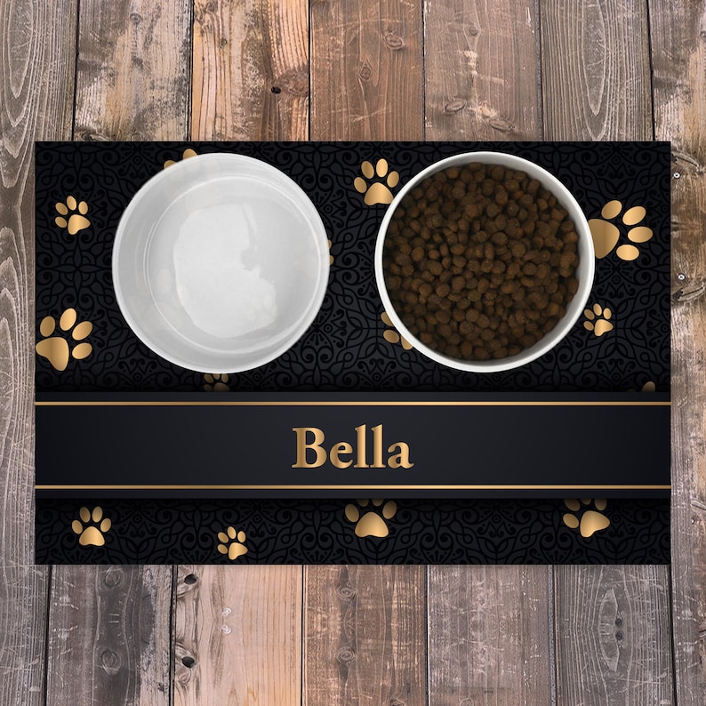 May include: A rectangular pet feeding mat featuring a black background with a dark ornate pattern and gold paw prints. A horizontal black band across the center displays the name Bella in gold text. Two white bowls sit on the mat, one empty and one filled with brown dry pet food.
