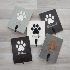 Puede incluir: Cinco ganchos de madera para correa de perro con huellas de patas. Los ganchos están pintados en diferentes colores, incluyendo gris, negro y blanco. Cada gancho tiene un nombre escrito en él: Amy, Belmont, Zarife, Miller y Mira.