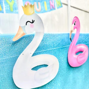 May include: A white swan-shaped pool float with a gold crown and a pink flamingo pool float on a blue surface. The swan has a light orange beak and a black curved eye. The flamingo is pink with a black beak.