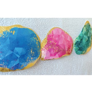 May include: Three abstract shapes with a gold foil finish. The shapes are blue, pink, and green and have a geometric pattern.
