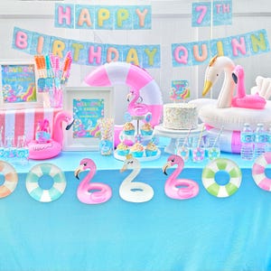 May include: A festive party scene with a blue tablecloth, featuring flamingo and pool float decorations. A banner reads "Happy Birthday Quinn" with a "7th" balloon. Cupcakes, a cake, and drinks complete the celebration.