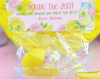 May include: A clear plastic bag filled with yellow lemon-shaped candies and a small lemon slice. The bag has a pink and yellow tag attached to it that says "You're The Zest! Thanks for making my party the best! Love. Quinn."
