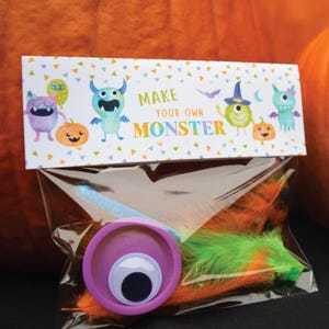 May include: A clear plastic bag filled with craft supplies for making a monster. The bag has a colorful label that says "Make Your Own Monster" with cartoon monsters and pumpkins. The supplies include feathers, pipe cleaners, googly eyes, and a purple play dough container.