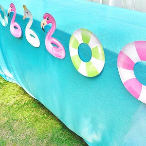 May include: A turquoise tablecloth is decorated with a garland of flamingo and life preserver cutouts. The flamingo cutouts are pink and white, and the life preservers are green and pink with white stripes. The scene is set on a green lawn.