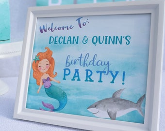 Op de afbeelding: Een wit ingelijst bord met een blauwe en groene aquarel achtergrond. Het bord zegt "Welkom bij: Declan & Quinn's birthday party!" met een illustratie van een zeemeermin en een haai.