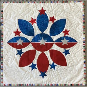 May include: A patriotic quilt featuring a central floral design in red and blue, with white star accents. The quilt has a white background with quilted patterns and a colorful border. The design is reminiscent of the American flag.