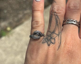Op de afbeelding: Een zilveren, met draad omwikkelde ring met een heldere, facetgeslepen steen wordt aan een vinger gedragen. De ring heeft een unieke, handgemaakte uitstraling. Een tweede zilveren ring met een decoratief patroon is ook zichtbaar.