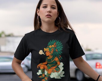 May include: Black t-shirt featuring a colorful illustration of an Aztec figure with a feathered headdress, sitting on a cloud. The shirt has the text "IKACHI ART Love for Our Roots" in the upper left corner. The model is wearing blue jeans.