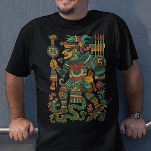 May include: Black t-shirt featuring a colorful illustration of an Aztec warrior holding a scepter and spear. The design includes a serpent, intricate patterns, and the text "Incaliart Love for Our Roots" in the upper left corner.