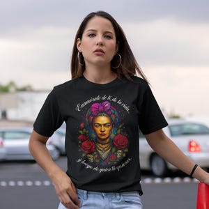 Könnte beinhalten: Schwarzes T-Shirt mit einem farbenfrohen Frida Kahlo-Porträt, verziert mit Rosen und Totenköpfen, und spanischem Text: "Enamórate de ti, de la vida... y luego de quien la quieras." Das Design beinhaltet ein farbenfrohes Kopftuch. Das T-Shirt wird mit blauen Jeans getragen.