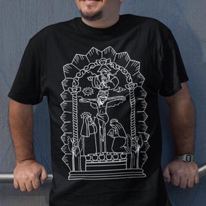 以下が含まれることがあります： 白い線画で宗教的なシーンを描いた黒いTシャツ。十字架にかけられた中心的な人物が描かれています。Tシャツはクルーネックで半袖です。「Inca Art Love For Our Roots」というテキストが含まれています。