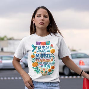 May include: White t-shirt with a colourful floral and skull design. The shirt features the text "SI NADA NOS SALVA DE LA MUERTE, QUE AL MENOS EL AMOR NOS SALVE DE LA VIDA". The shirt is paired with blue jeans.