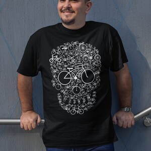 May include: A black t-shirt with a white design of a skull made up of bicycles. The person wearing the shirt is standing in front of a blue wall.