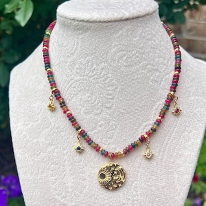 May include: A beaded necklace featuring small, multi-colored gemstone rondelles in shades of red, green, and yellow. The necklace includes four gold-toned charms: three small star-shaped pendants and one larger circular pendant depicting a sun and moon.